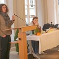 Eine Frau steht an einem Rednerpult, daneben sitzt eine Frau an einem Tisch