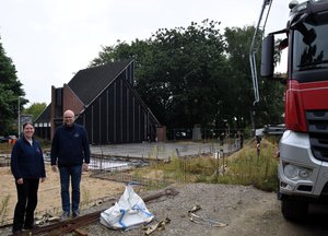 Eine Frau und ein Mann stehen links, rechts ein Teil eines roten LKW. Mittig eine Baustelle.