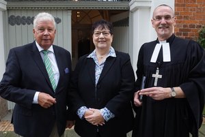 Zwei Männer und eine Frau in dunkler Kleidung stehen vor einer offenen Kirchentür. Einer der Männer trägt Talar und ein Kreuz um den Hals.