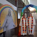 Zwei Frauen in weißer geistlicher Kleidung mit regenbogenbunten Schal stehen unter einem regenbogenbunten Bogen