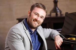 Ein Mann mit Brille und Sakko sitzt an einem Klavier und lächelt in die Kamera