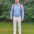 Ein Mann in heller Hose und blauem Hemd sowie roten Hosenträgern sowie einer Brille lächelt in die Kamera