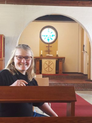 Eine blonde Frau mit Brille sitzt in einer Kirchenbank und lächelt in die Kamera, im Hintergrund ein Altar und ein Kirchenfenster