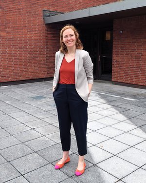 Eine Frau mit halblangen Haaren lächelt in die Kamera, im Hintergrund ein Gebäude