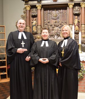 Ein Mann und zwei Frauen in geistlicher Kleidung stehen vor einem Altar und lächeln in die Kamera