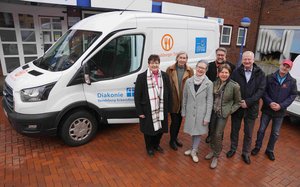 Sieben Menschen stehen vor einem Transporter mit aufgedruckten Logos und offener Fahrertür