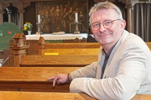 Ein Mann mit Brille in hellem Anzug sitzt in einer Kirchenbank und lächelt in die Kamera