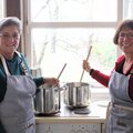 Zwei Frauen in Schürzen stehen vor großen Kochtöpfen und halten Kochlöffel in der Hand