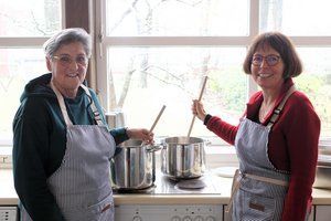 Zwei Frauen in Schürzen stehen vor großen Kochtöpfen und halten Kochlöffel in der Hand