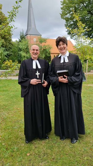 Propst Krüger und Pastorin Andersson stehen auf einer Wiese, im Hintergrund die Hohner Kirche