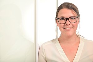 Eine junge Frau in heller Kleidung, mit Brille und braunen Haaren schaut in die Kamera