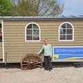 Eine Frau steht im Sand vor einem Holzbau auf Rädern mit der Aufschrift "Schäferwagenkirche"
