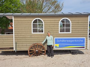 Eine Frau steht im Sand vor einem Holzbau auf Rädern mit der Aufschrift "Schäferwagenkirche"