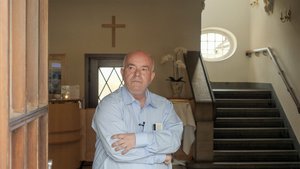 Herr Zander steht in einem hellen Hemd in einem Treppenhaus, die Arme vor der Brust verschränkt, im Hintergrund ein Kreuz an der Wand.