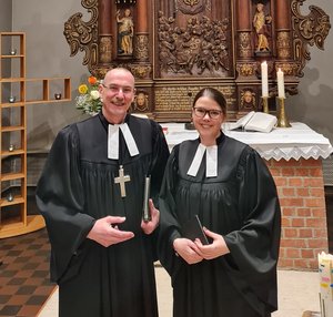 Ein Mann und eine junge Frau in geistlicher Kleidung stehen in einer Kirche und lächeln in die Kamera
