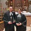 Ein Mann und eine junge Frau in geistlicher Kleidung stehen in einer Kirche und lächeln in die Kamera