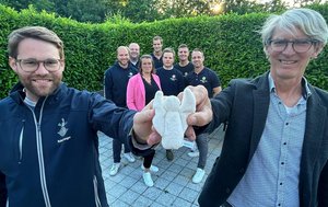 Zwei Männer halten einen kleinen Stoffengel in die Kamera, im Hintergrund eine Gruppe von sieben Menschen