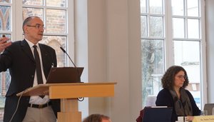 Ein Mann steht am Rednerpult, daneben sitzt eine Frau an eine, Tisch.