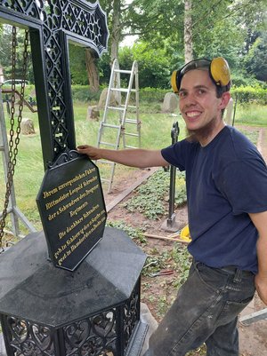 Junger Mann in Arbeitskleidung lächelt in die Kamera, hält schwarze Platte mit goldener Schrift