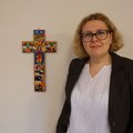Eine junge Frau mit lockigen Haaren und einer Brille steht neben einem bunten Kreuz, welches an der Wand hängt