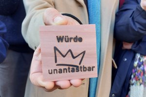 Eine Hand hält ein Holzstück mit der Aufschrift "Würde unantastbar" und einer Krone darauf