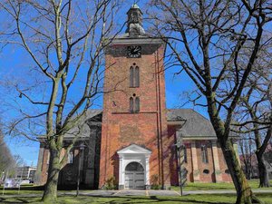 Ein Kirchgebäude im Sonnenschein mit Blick auf das Hauptportal, davor stehen Bäume noch ohne Laub.