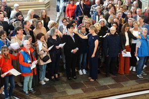 Eine Gruppe von Menschen steht im Altarraum einer Kirche. In der Mitte wird eine Frau gesegnet. Die Menschen legen jeweils eine Hand auf die Schulter ihrer Nachbarn.