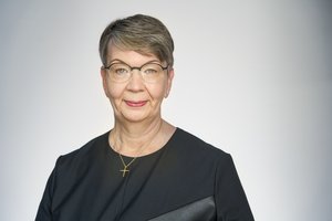 Eine Frau mit Brille und kurzen dunklen Haaren steht vor einem grauen Hintergrund und schaut in die Kamera
