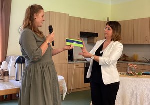 Eine Frau mit Mikro in der Hand überreicht einer weiteren Frau einen Scheck