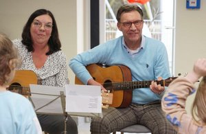 Ein Mann und eine Frau sitzen mit Gitarren vor einigen Kindern
