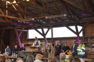 Ein Mann am Mikrofon, daneben mehrere andere Menschen, im Vordergrund viele sitzende Menschen, die zuhören