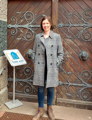 Eine Frau steht im Mantel vor einer Kirche, daneben ein Schild "Die Kirche ist offen"