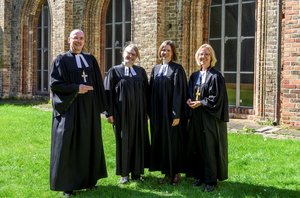 Ein Mann und drei Frauen in geistlicher Kleidung stehen im Sonnenschein auf einer Wiese, im Hintergrund eine Kirche.