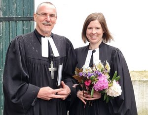 Ein Mann und eine Frau in geistlicher Kleidung (Talar) stehen nebeneinander. Die Frau hält einen Blumenstrauß.