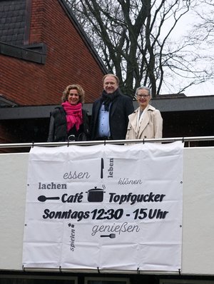 Zwei Frauen und ein Mann stehen hinter einem Banner mit der Aufschrift "Café Topfgucker"