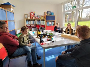 Ein Mann mit Kind auf dem Schoß im Gespräch mit Jugendlichen, alle sitzen an einem Tisch, auf dem Holz und Spielzeugfahrzeuge liegen