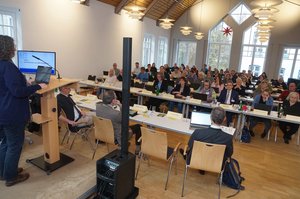 Eine Frau am rechten Rand des Bildes steht am Rednerpult und schaut in eine Gruppe an Menschen, die an Tischen sitzen.