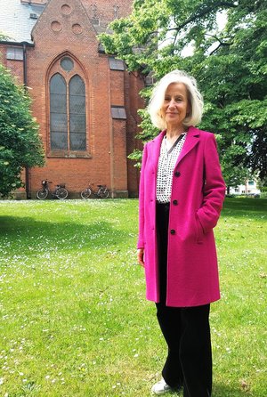 Eine Frau im pinken Mantel steht auf einer Wiese, im Hintergrund ein Kirchgebäude