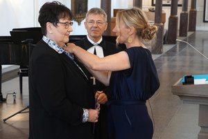 Das Bild zeit zwei Frauen und einen Mann im Talar mit Kreuz um den Hals. Eine jüngere blonde Frau heftet einer Frau mit dunklen Haaren etwas an die Kleidung.