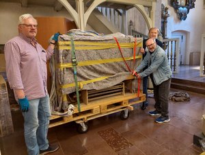 Drei Männer stehen in einem Kirchraum, in der Mitte ein verpackter Kasten