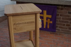 Ein Holzkasten mit der Aufschrift "Gebetskasten", im Hintergrund ein lila Tuch mit Kreuzen darauf