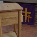 Ein Holzkasten mit der Aufschrift "Gebetskasten", im Hintergrund ein lila Tuch mit Kreuzen darauf