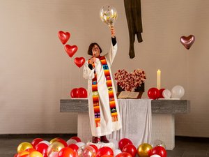 Eine Frau im weißen Talar mit regenbogenfarbiger Stola, im Vordergrund viele herzförmige, rote und rosige Luftballons