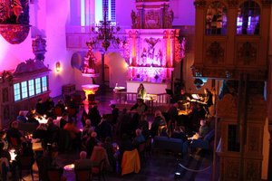 Viele Menschen sitzen an Tischen im Altarraum einer Kirche, der pink beleuchtet ist